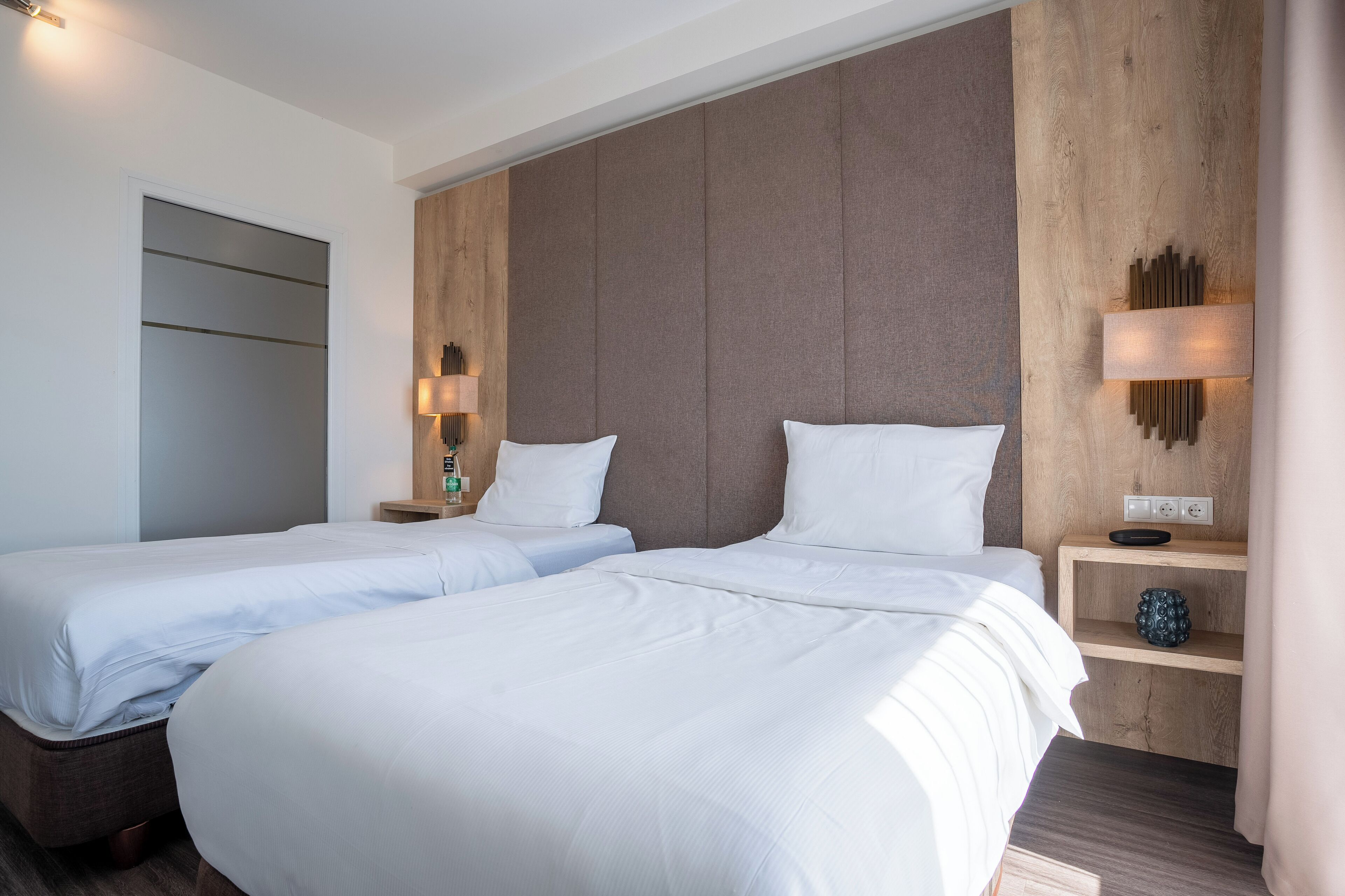 Habitación de hotel con dos camas, cabecero de madera, mesitas de noche con lámparas y un estante pequeño con un objeto decorativo.