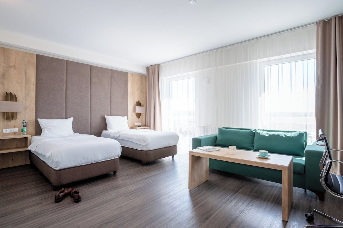 Een hotelkamer met twee bedden, een groene bank, een houten salontafel en uitzicht op de stad door grote ramen.