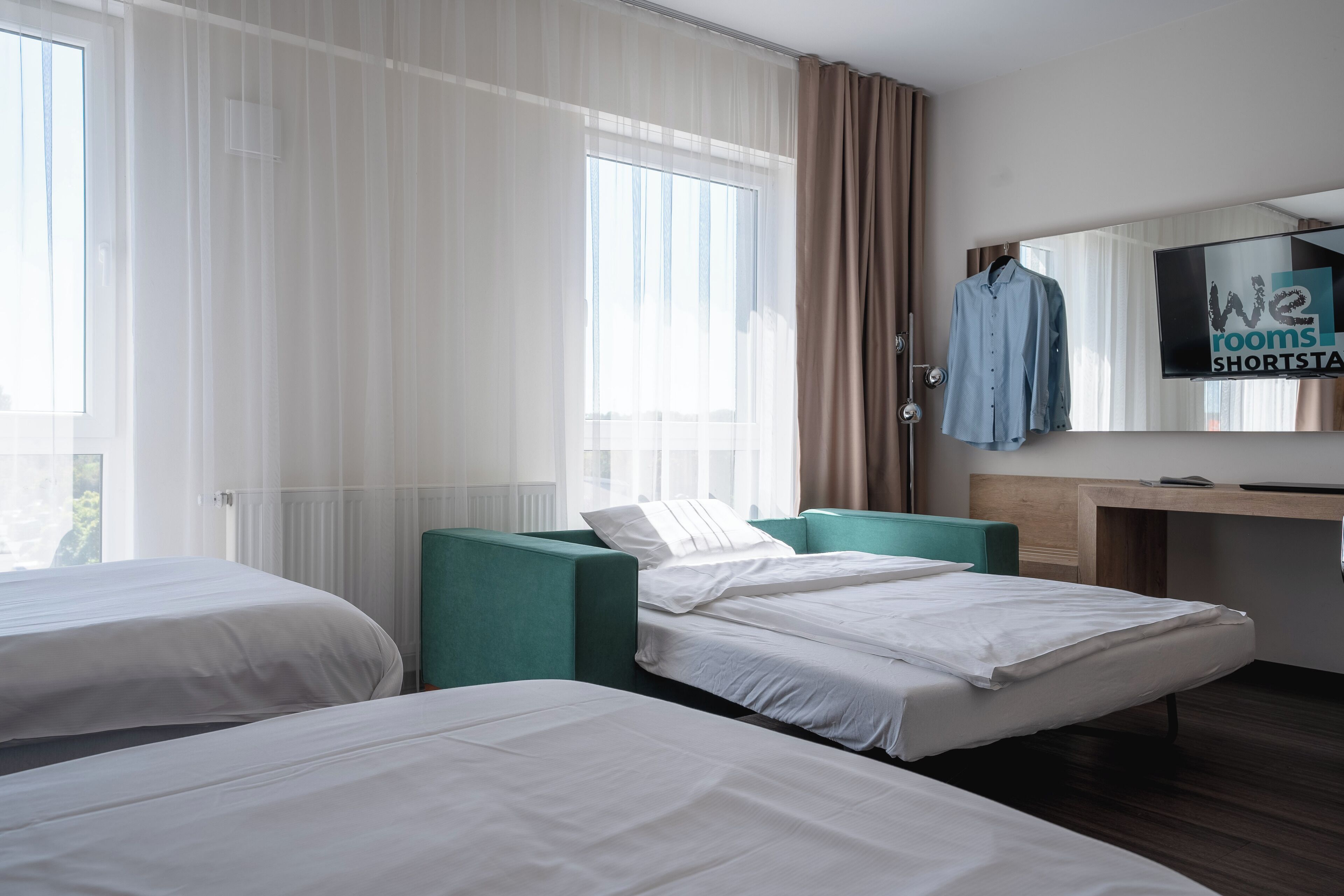 A hotel room with two single beds, a desk, a chair, a mirror, and a window with curtains.