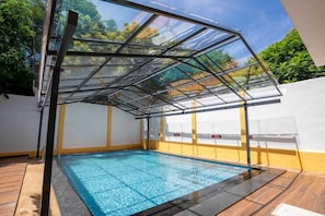 Una piscina con cerramiento de vidrio, rodeada por un muro blanco y amarillo, y una zona de terraza de madera.