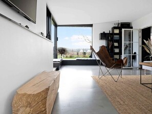 A modern living room with a large window, a wooden bench, a folding chair, and a bookshelf. - 5 Star Holiday Home in Aakirkeby (Akirkeby)