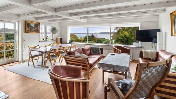 Amplia sala de estar con una mesa de comedor, varias sillas, un sofá y una televisión. Amplias ventanas ofrecen vistas al exterior.
