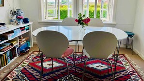 Een eetgedeelte met een ronde tafel, vier stoelen, een vaas met bloemen en een boekenkast met boeken en decoratieve objecten.