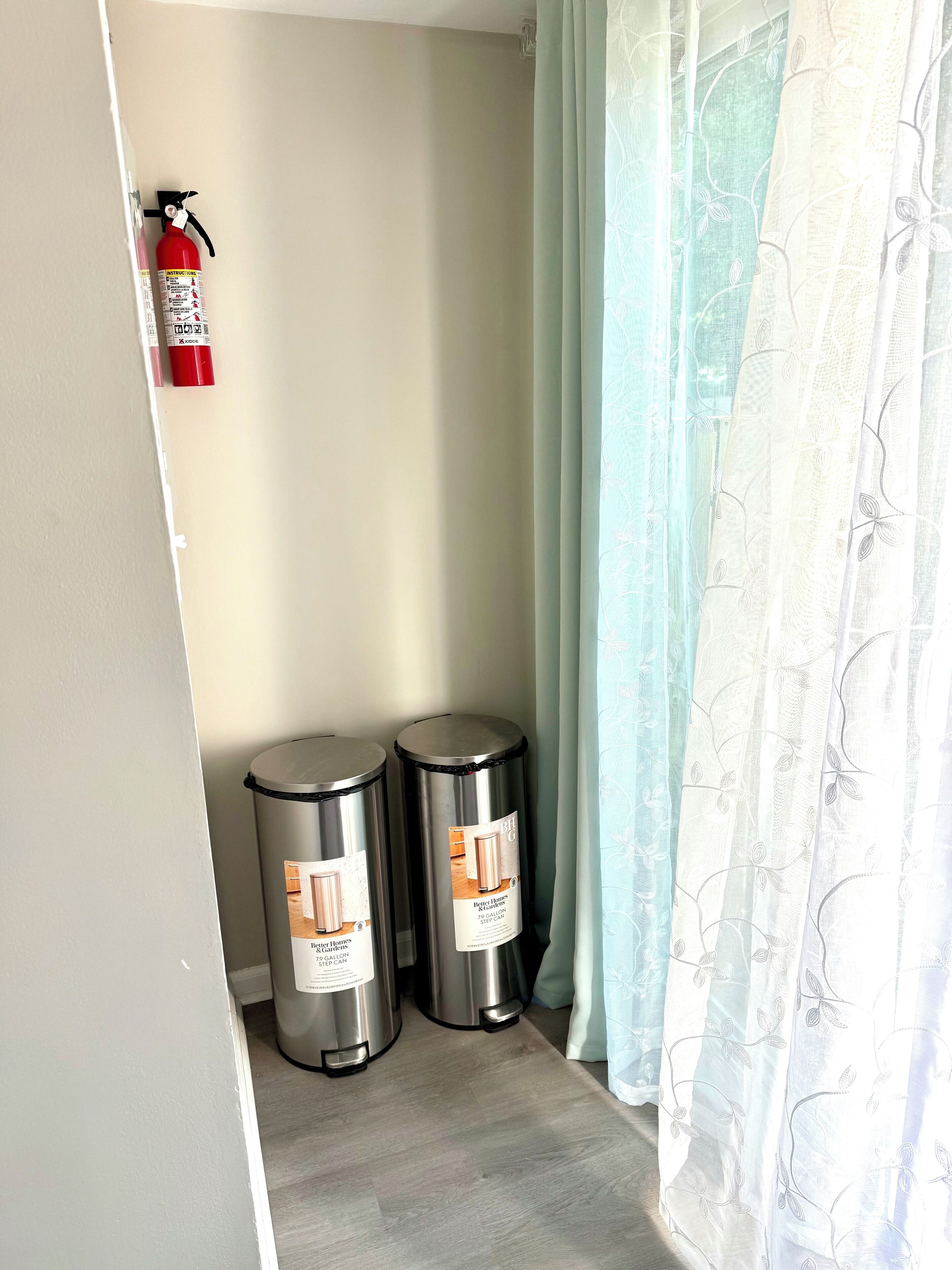 A corner of a room with a fire extinguisher mounted on the wall, two trash bins, and a sheer curtain.