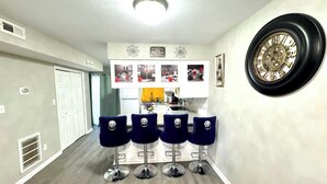 Un couloir avec un coin cuisine, une horloge au mur et quatre tabourets de bar bleus.