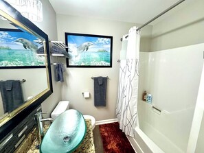 Ein Badezimmer mit einem Glaswaschbecken, einer Toilette, einer Dusche mit weißem Vorhang und einem gerahmten Bild von Delfinen an der Wand.