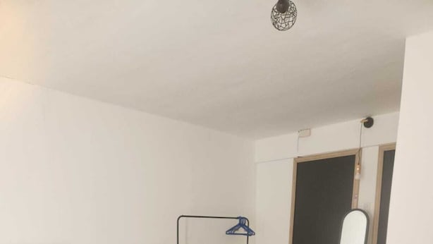 A bedroom with a bed, a bedside table, a hanging clothes rack, and a door leading to another room.