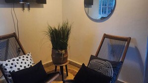 A living room with two wooden chairs, a small potted plant, a round mirror, and a flat-screen TV mounted on the wall.