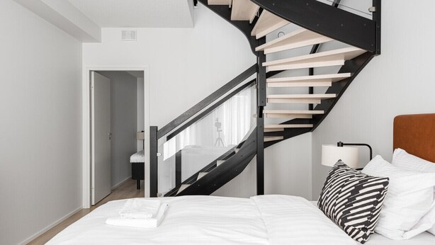 A modern bedroom with a curved black staircase, a bed with white bedding, and a nightstand with a lamp. - Borg Loft Apt Itäpuisto (Pori)