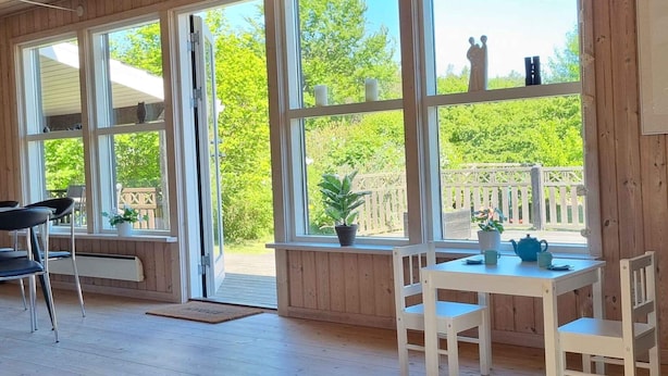 A room with large windows, wooden walls, a white table with chairs, and a view of a wooden deck and greenery outside. - 5 Star Holiday Home in Ebeltoft (Ebeltoft)
