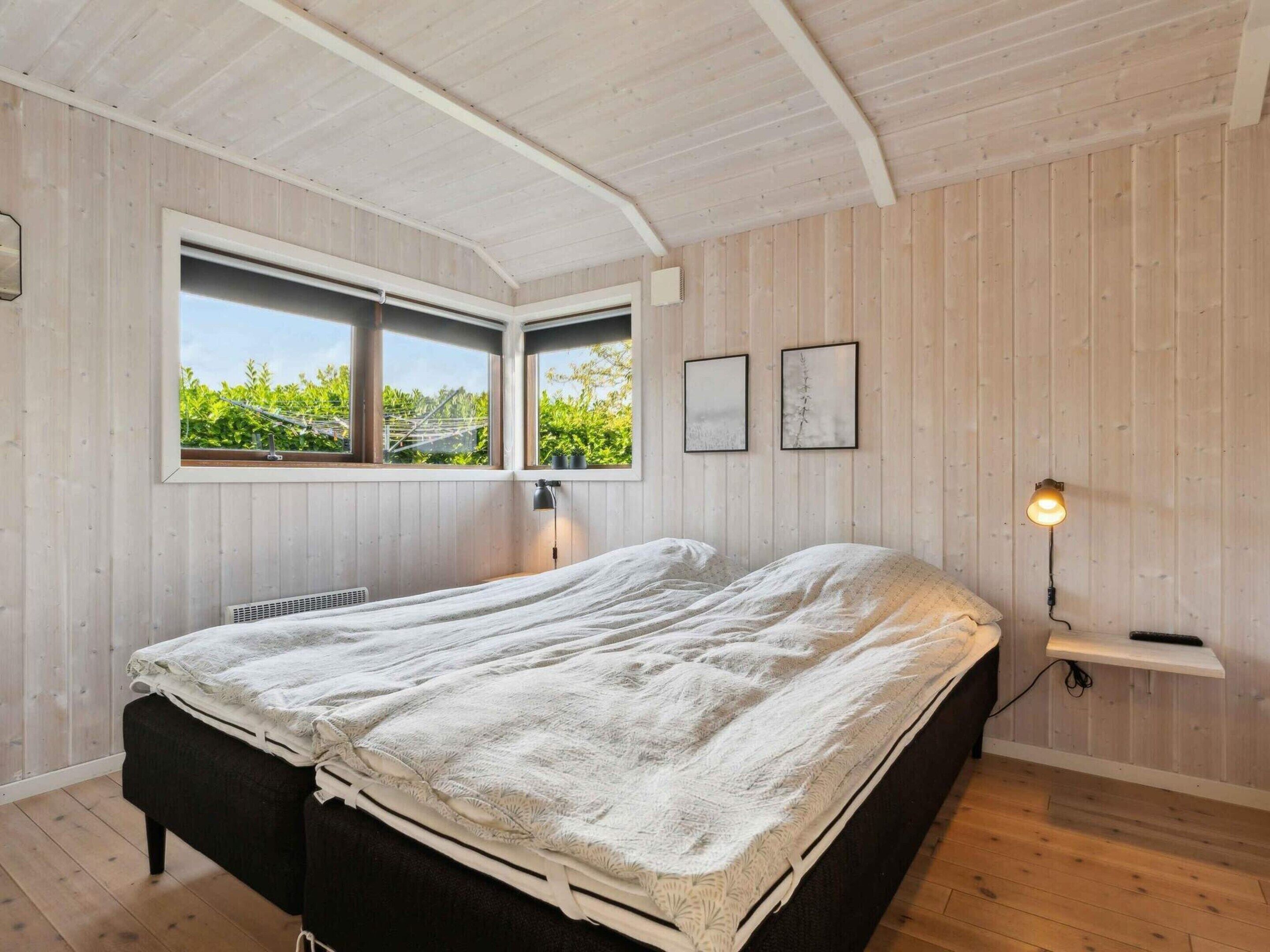 A bedroom with a bed, two windows, a radiator, a wall-mounted shelf, and a wall-mounted lamp.