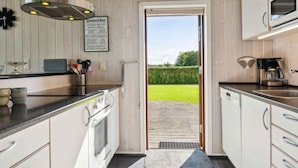 Una cucina moderna con mobili bianchi, forno integrato, microonde e macchina da caffè. Un cartello è appeso al muro e si può vedere un giardino attraverso una porta aperta.