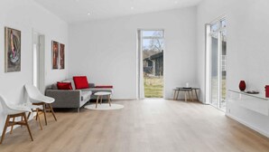 Un salon spacieux avec un canapé gris, des chaises blanches, une petite table ronde et une grande fenêtre donnant sur l’extérieur.