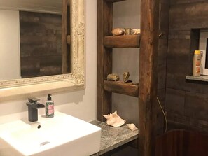 Une salle de bain avec un lavabo blanc, un grand miroir et des étagères en bois où sont rangés les articles de toilette et des objets décoratifs.