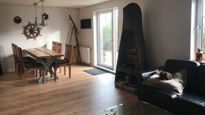 A living room with a wooden dining table, chairs, a black sofa, a fireplace, and a large window. - 5 Star Holiday Home in Farso (Farso)