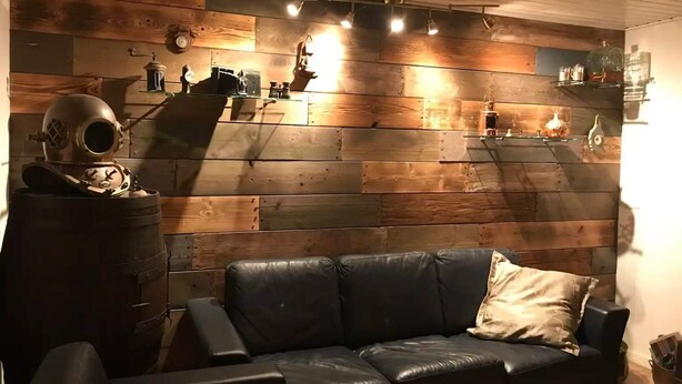 A room with a wooden wall, a black leather sofa, and a vintage helmet on a barrel. - 5 Star Holiday Home in Farso (Farso)