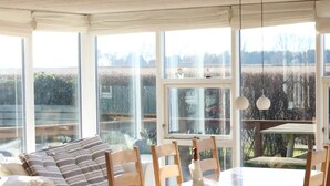 Ein heller Essbereich mit großem Tisch und Stühlen, großen Fenstern mit Blick ins Freie und einer gemütlichen Sitzecke mit Sofa und Kissen.
