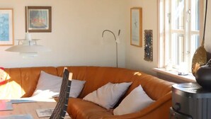 Ein gemütliches Wohnzimmer mit einem braunen Ledersofa, einem Holztisch und einem schwarzen Sessel. Ein Fenster mit Vorhängen, eine Stehlampe und ein dekoratives Wandbild sind ebenfalls zu sehen.
