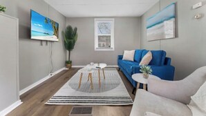 Un salon moderne avec un canapé bleu, un fauteuil blanc et une table basse. Un téléviseur est fixé au mur, et une plante en pot est posée à proximité.