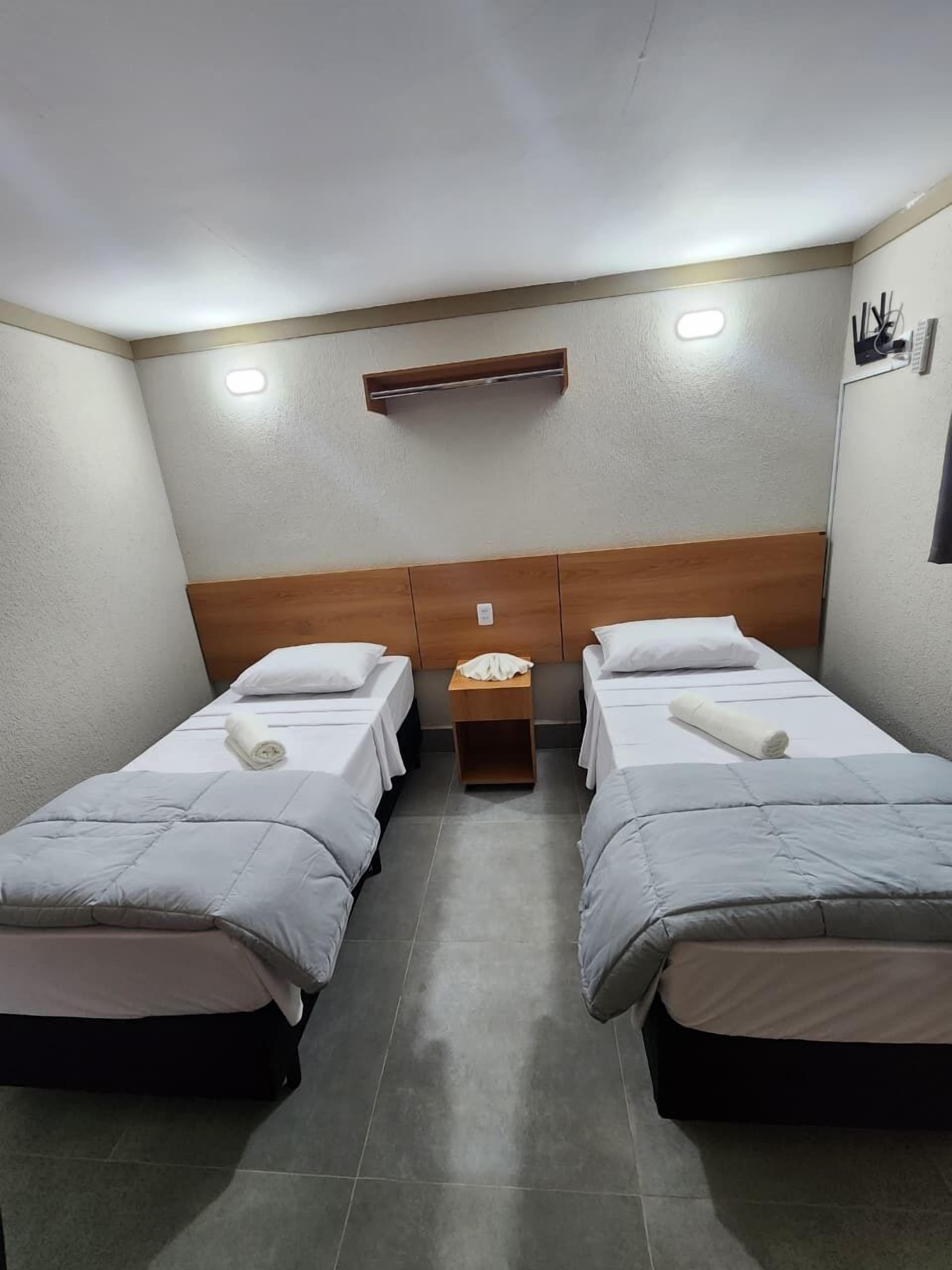 A hotel room with two single beds, a wooden headboard, a small bedside table, and a wall-mounted shelf.