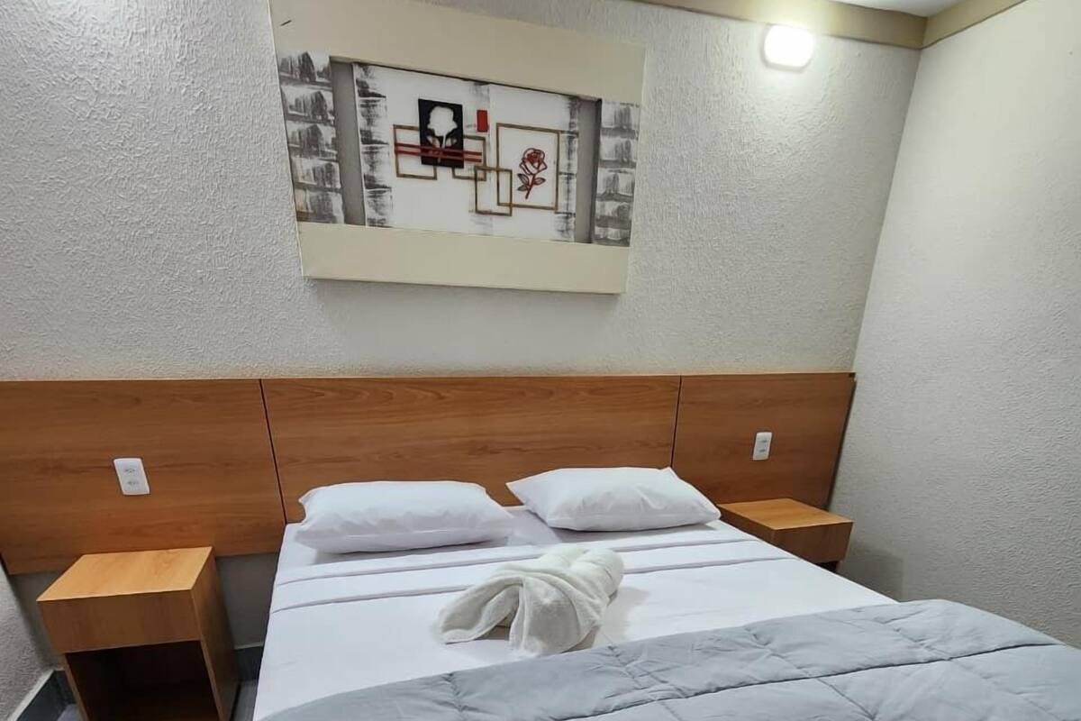 Un lit bien fait, avec une couette grise, deux oreillers blancs et une serviette blanche pliée. On y trouve deux tables de chevet en bois avec des lampes, et une œuvre d’art murale au-dessus du lit.