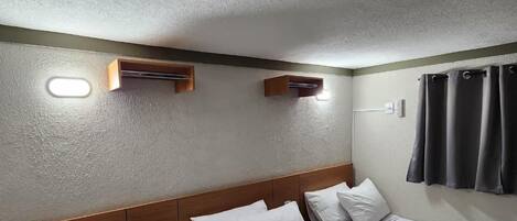 Una habitación de hotel con tres camas individuales, un cabecero de madera, un pequeño estante de madera y una lámpara fijada a la pared.