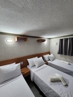 Una habitación de hotel con tres camas individuales, un cabecero de madera, un pequeño estante de madera y una lámpara fijada a la pared.