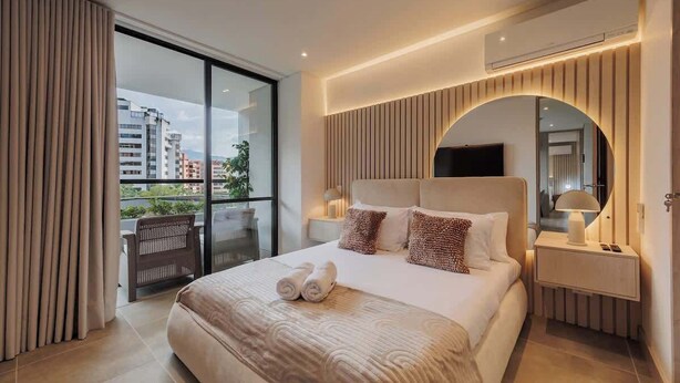 A modern hotel room with a large bed, a TV, a bedside table, and a view of the city through a large window.