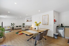 A modern living room with a dining table, chairs, a sofa, and a kitchen area with a dishwasher.