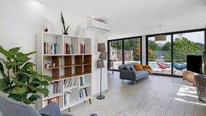 En moderne stue med bogreol, grå sofa og stort vindue med udsigt over en pool.