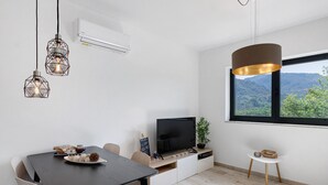 Una sala de estar moderna con televisor, mesa de comedor, sillas, lámparas colgantes y un amplio ventanal con vistas.