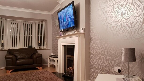 A living room with a fireplace, a television mounted above it, a brown leather sofa, and a patterned wallpaper. - Charming Liverpool Home With Gardens and Parking (Liverpool)