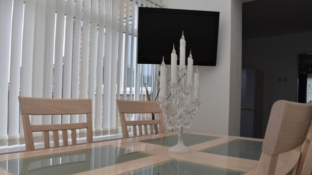 A dining area with a glass-top table, two wooden chairs, a white candelabra, and a flat-screen TV mounted on the wall. - Charming Liverpool Home With Gardens and Parking (Liverpool)