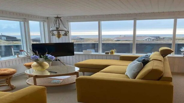 Una sala de estar con un sofá amarillo, un televisor sobre un mueble bajo, una mesa redonda con un jarrón y ventanas grandes que ofrecen vistas del paisaje.