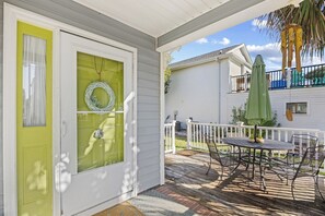 Een overdekt terras met een tafel en stoelen, een groene deur en een witte hek.