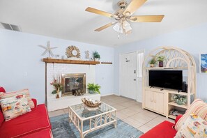 Een woonkamer met een open haard, rode bank, glazen salontafel, plafondventilator en een televisie op een houten dressoir.