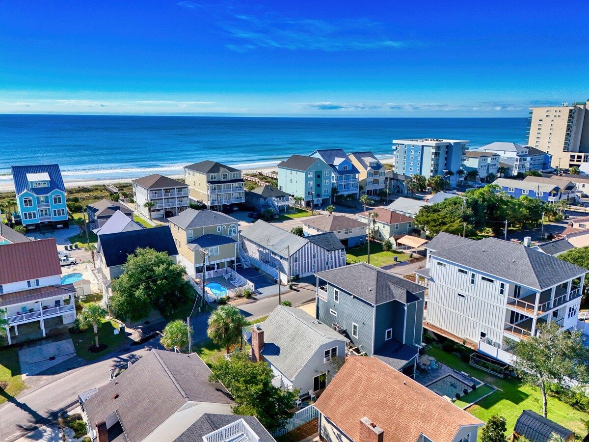 Un quartier résidentiel situé sur la côte, avec de nombreuses maisons à deux étages, une plage en arrière-plan et un ciel dégagé.