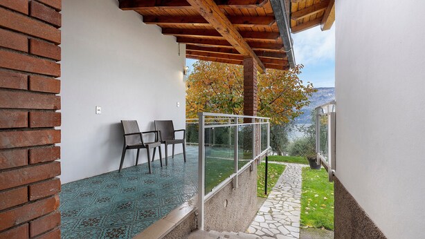 A covered outdoor area with a brick wall, a glass railing, and a view of a garden and mountains. - Apartment 'Bungalow Pit Stop' with Lake View, Wi-Fi and Air Conditioning (Brenzone sul Garda)