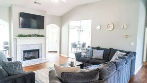 A living room with a fireplace, a sofa, a TV, and a dining area in the background.