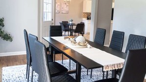A dining area with a long table, six chairs, a chandelier, and a kitchen in the background.