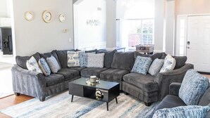 A living room with a sectional sofa, a coffee table, and a rug. There are three clocks on the wall and a door to the right.
