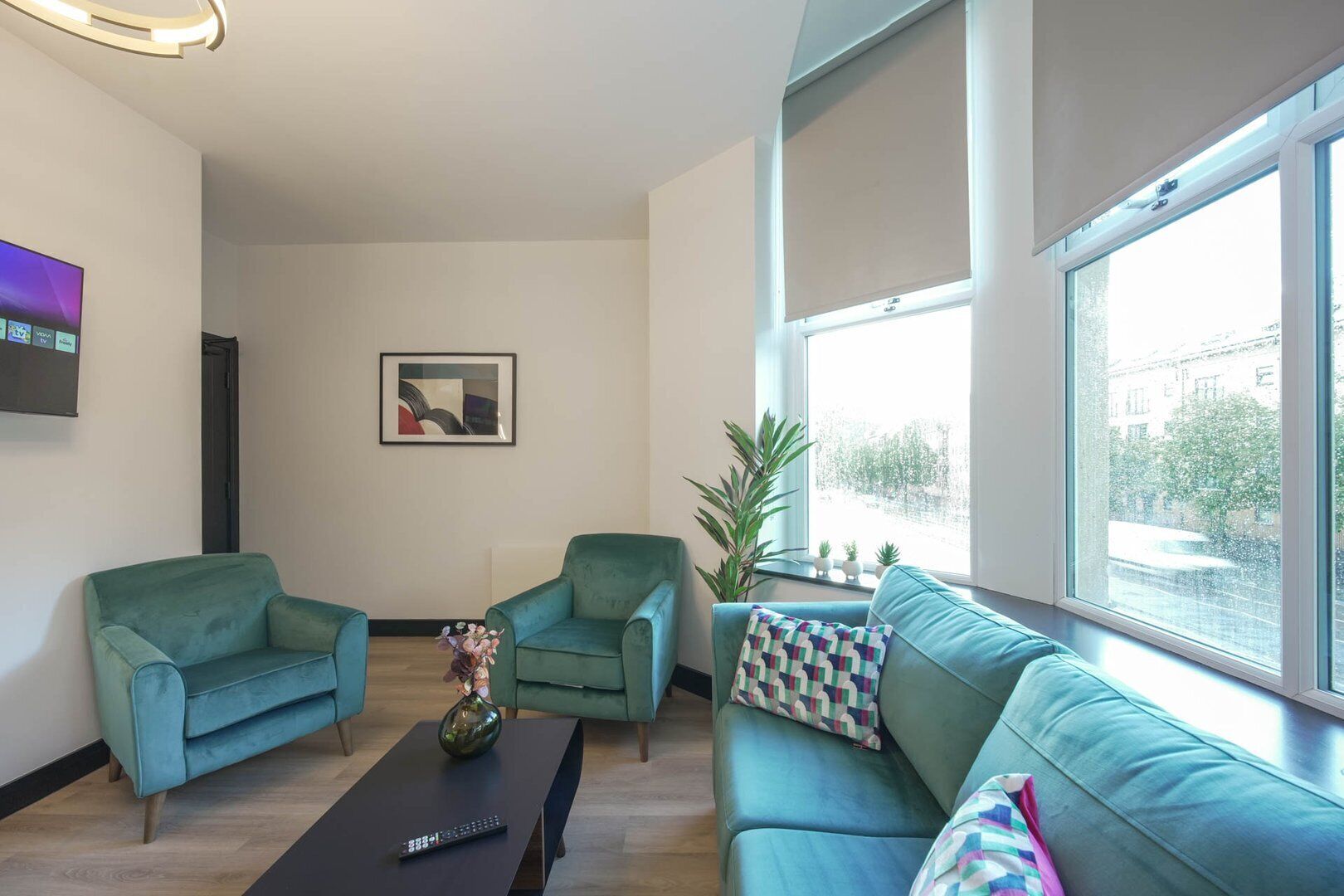 A modern living room with two teal sofas, a coffee table with a vase, and a flat-screen TV on the wall.