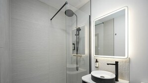 Une salle de bain moderne dotée d’une douche à parois de verre, d’un lavabo rond avec un receveur noir et d’un miroir dont le cadre est illuminé.