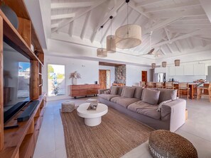 A spacious living room with a large TV, a sofa, a coffee table, and a dining area with chairs and a kitchen in the background. - Villa Surfside (Sainte-Anne)