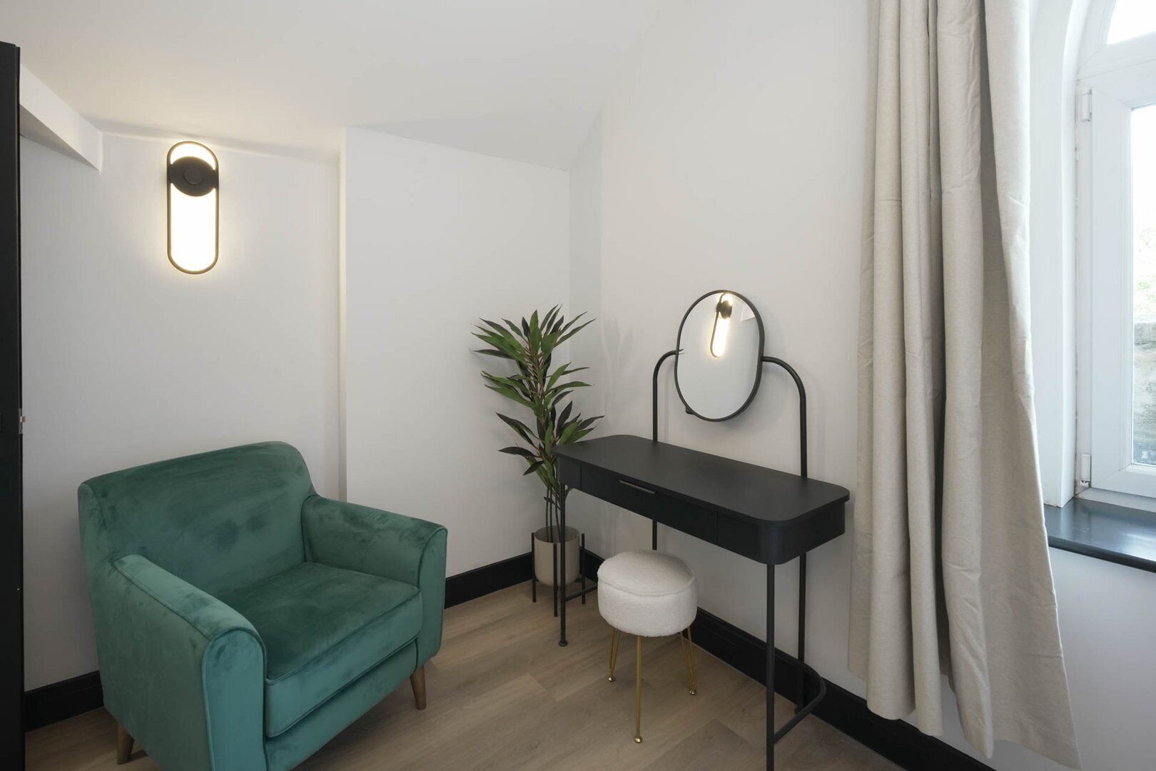 Una habitación con un sillón turquesa, un taburete blanco pequeño, un tocador negro con espejo y una planta en maceta.