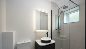 Ein modernes Badezimmer mit einer gläsernen Duschkabine, einem runden Waschbecken mit Spiegel darüber und einer Toilette links.