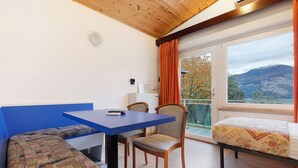 Ein Zimmer mit blauem Tisch, zwei Stühlen, einer Bank, einem Bett mit floralem Überwurf und einem Fenster mit orangefarbenen Vorhängen sowie Bergblick.