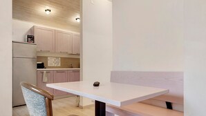 Eine kleine Küchenzeile mit Kühlschrank, Mikrowelle und Spüle. Ein weißer Tisch wird von einer Bank und einem Stuhl mit Korbsitz ergänzt.