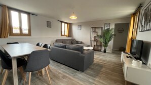 Una sala de estar moderna con un sofá gris, una mesa de comedor con sillas, un mueble para la televisión y una planta en maceta.