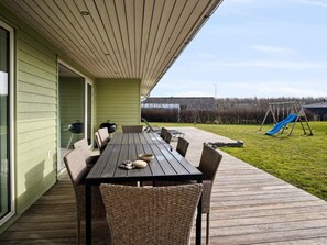Une terrasse couverte avec une table en bois et des chaises en osier, un barbecue et une aire de jeux avec un toboggan.